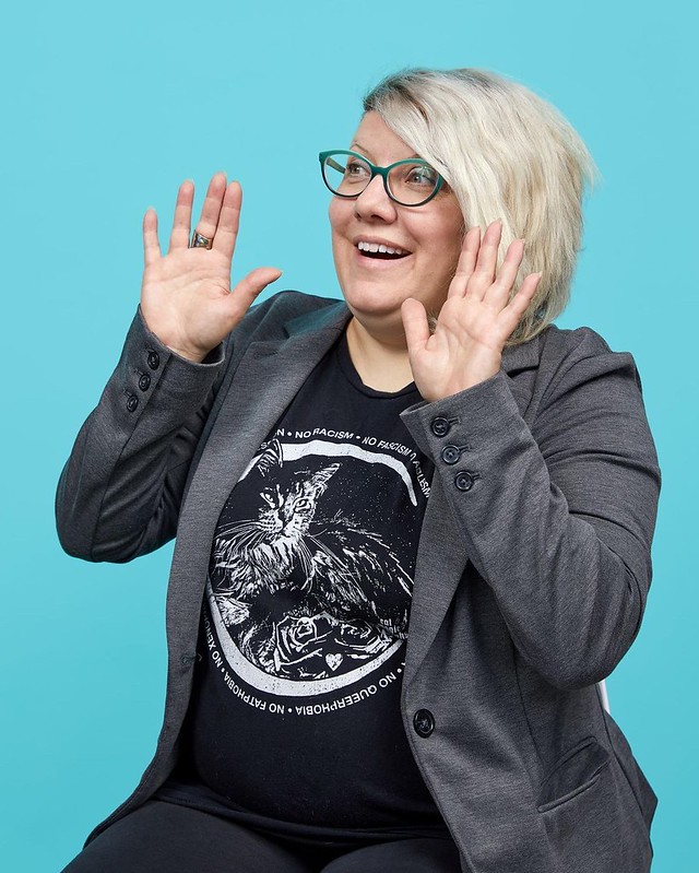 Sharyn sitting in front of a teal background holding their hands up as though they're shouting