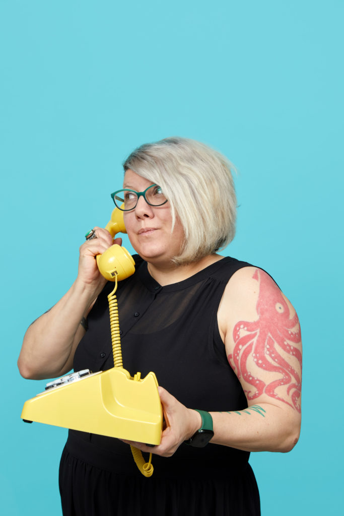 3/4 view of Sharyn wearing glasses while pretending to talk on a yellow landline telephone handset