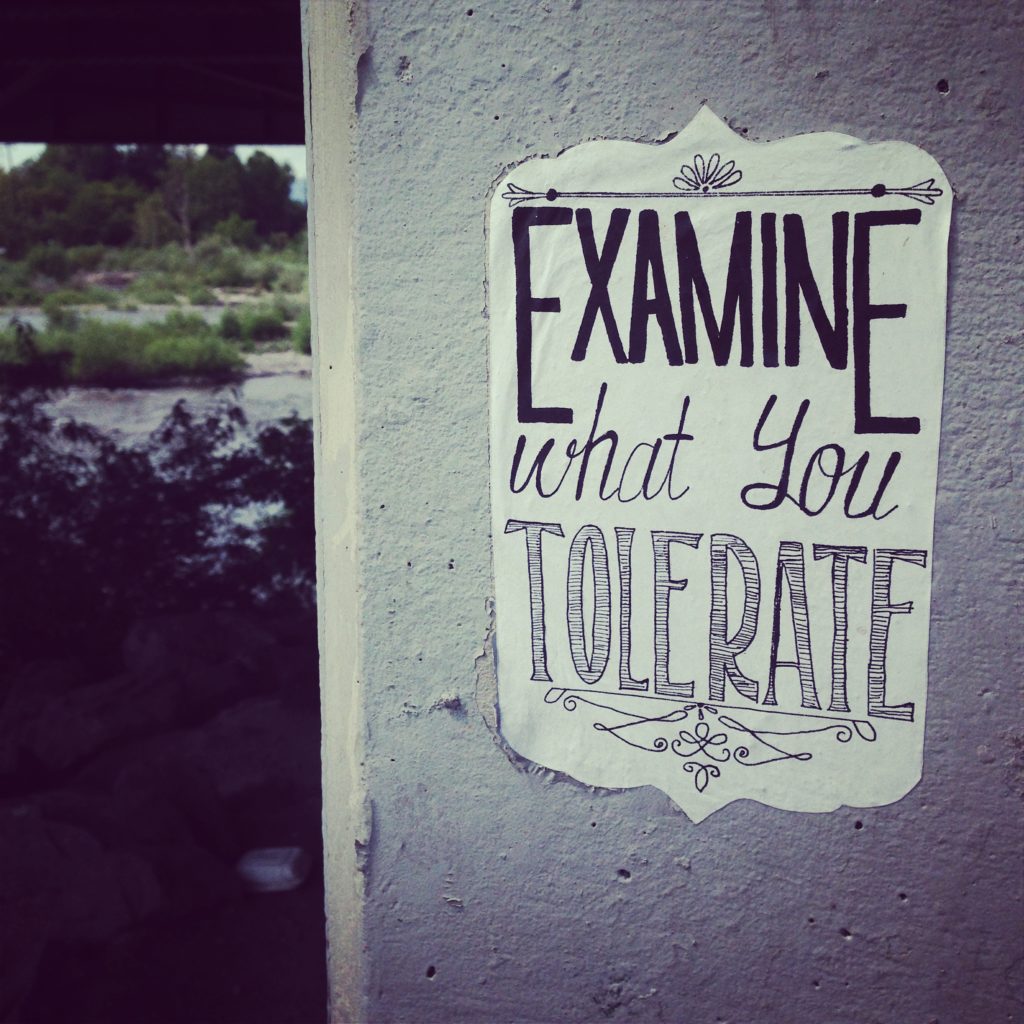 A stone wall with a paper sign affixed to it with the text Examine what you tolerate