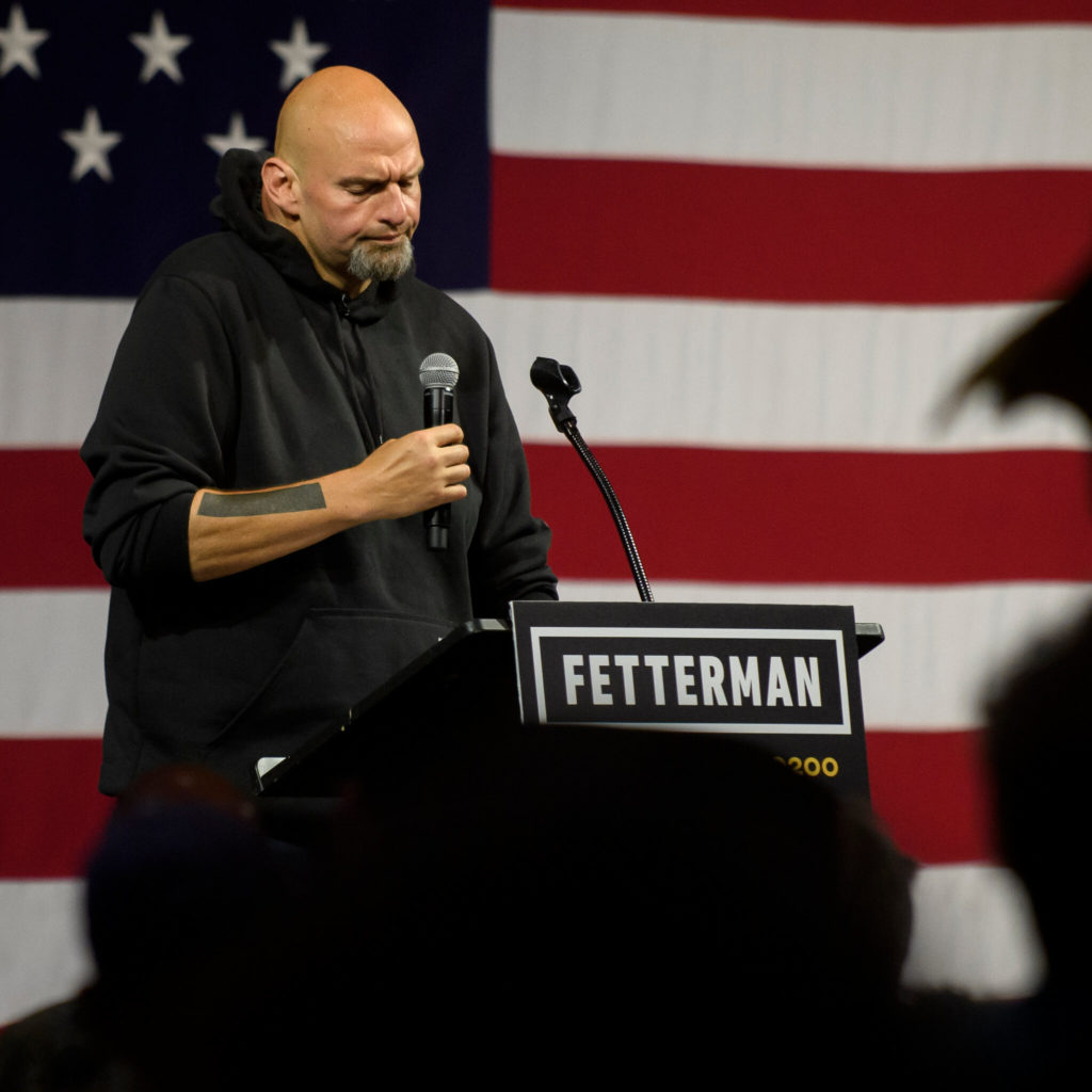 Politician John Fetterman looking thoughtful behind a podium, with a microphone in hand, and a large American flag as a backdrop