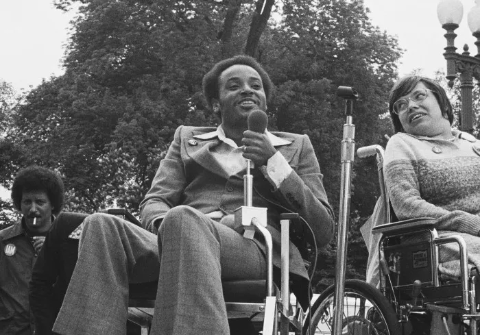 Black and white photo from 1977 of disability activist Brad Lomax holding a microphone with activist Judith Heumann sitting next to him, both in their wheelchairs.