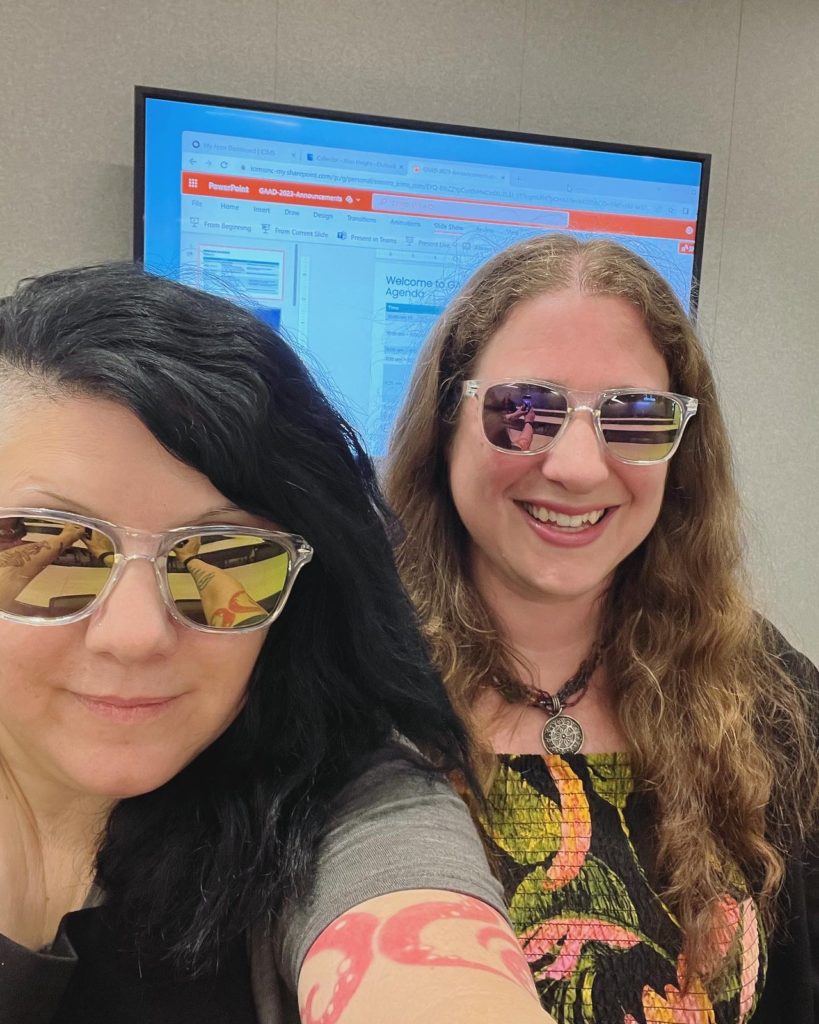 Two accessibility team members wearing icims branded sunglasses in a conference room with a screen behind them showing a powerpoint slide deck