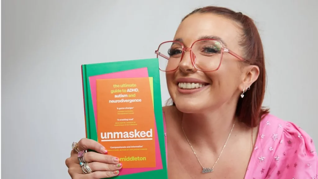 Author Ellie Middleton grinning while holding a copy of her book Unmasked: the ultimate guide to ADHS, autism, and neurodivergence
