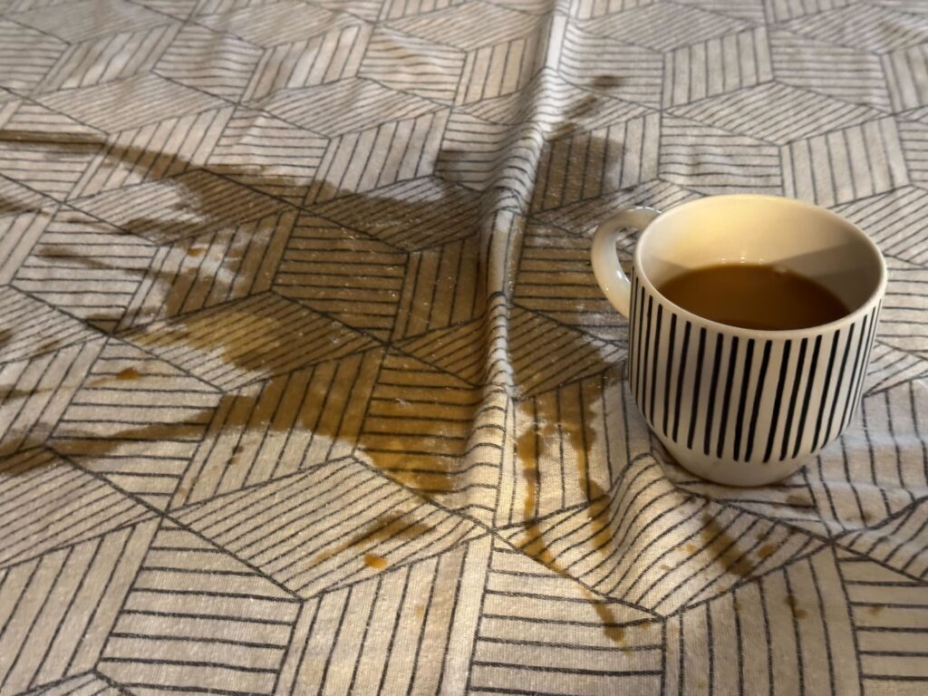 A black and white mug sitting on a table with a black and white fabric tablecloth. A large amount of the coffee in the mug has been spilled onto the tablecloth.