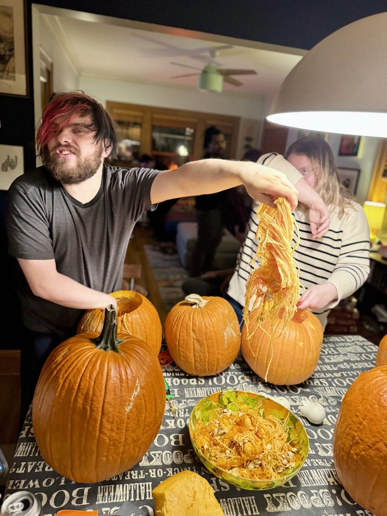 A young bearded man at a pumpkin carving party scooping out pumpkin guts into a bowl